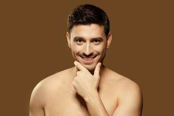 Close-up portrait of young man standing isolated on brown background, looking at camera with cunning smile and holding his chin as if flirting. Men beauty skin care ads
