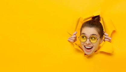 Fototapeta Horizontal banner of young girl in glasses tearing paper and peeking out hole, curious about commercial offer on copy space on left, isolated on yellow background obraz