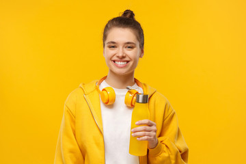 Happy girl in t-shirt and hoodie, wearing wireless headphones around neck, holding reusable water...