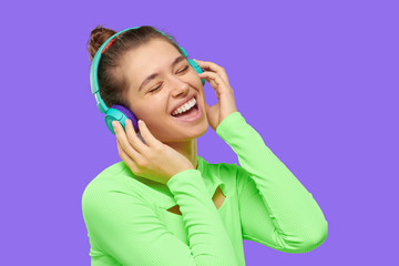 Young happy laughing girl listening to music in wireless headphones with closed eyes, dressed in neon green top, having fun, isolated on purple background