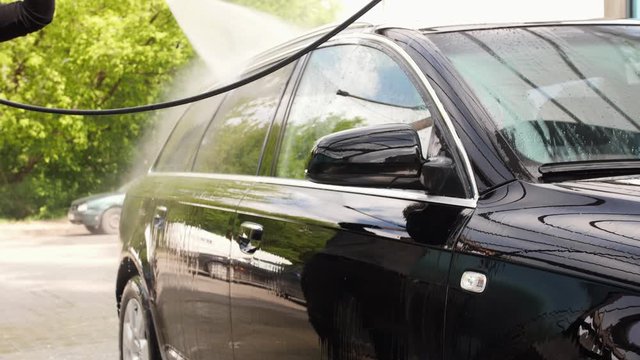 A man is rinsing a black car at a self-wash car point. He is using the spray wand and the water is going through it with high pressure. This is a cheap way to do a good clean of a car.