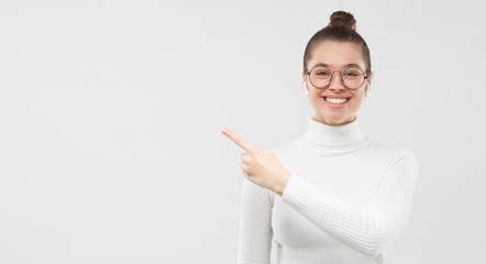 Horizontal banner of girl in eyeglasses and white sweater, smiling to camera, pointing to copy space on left side, isolated on gray background