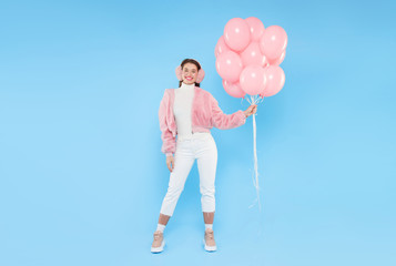 Young brunette girl wearing pink furry bomber and earmuffs, standing with bunch of pink balloons, celebrating her birthday, isolated on blue background