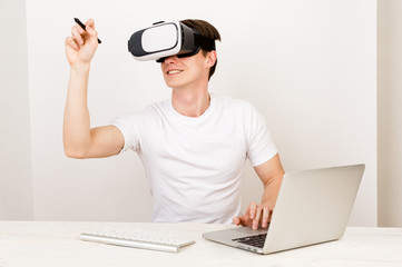Male using virtual reality headset interacts with augmented things orienting in three dimensional space while sitting on his desk with keyboard and laptop on white background.