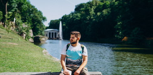 Sofia Park, Uman. Handsome man with a backpack sitting on a stone bench in the park. Young man on the background of the lake with a tall fountain. A man with a backpack on a tour of a national park.
