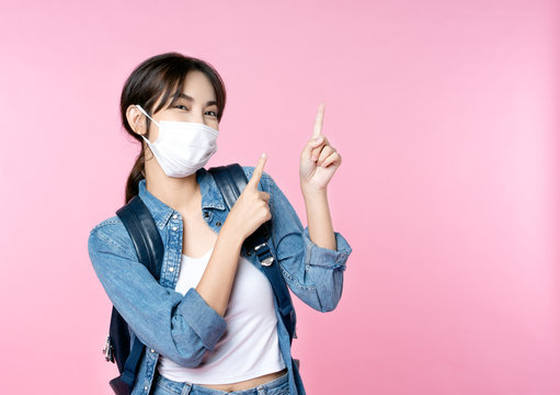 Young happy Asian woman pointing finger hands up to copy space wearing hygienic mask to prevent infection corona virus she jean jacket and carry backpack shirt shoot in isolated on pink background