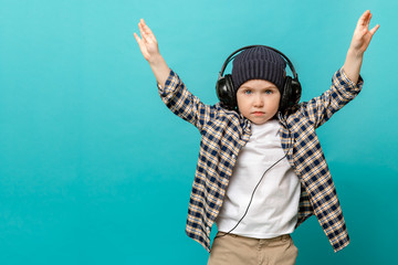 A fashionable 4-5-year-old kid in a checked shirt and a white t-shirt with a hat listens to music with headphones and raises his hands up, standing out against the pastel blue background of a children