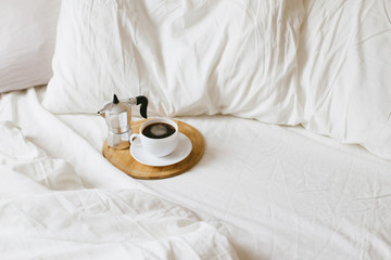 Cozy bedroom details: cup of hot fresh coffee and coffeemaker on wooden tray in bed with white...