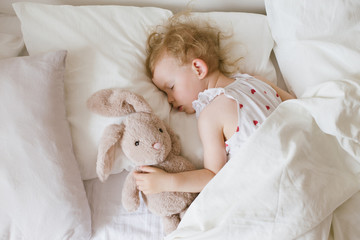 Cute child little girl sleeping in bed with white linen hugging her soft toy teddy bunny.