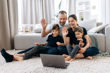 Video call. A happy family, young parents and their two adorable daughters communicate via video conference with friends sitting at home on the floor. Online communication