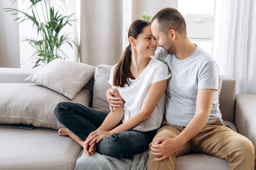 Beautiful couple in love in stylish casual wear hugging while sitting on a sofa at home. Attractive caucasian girl and handsome guy in love