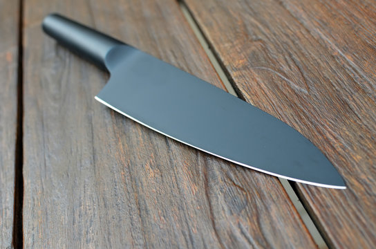 Close-up of a kitchen knife on wooden background with a black handle and black blade, matte finishing, flat lying, perspective view, focus on foreground