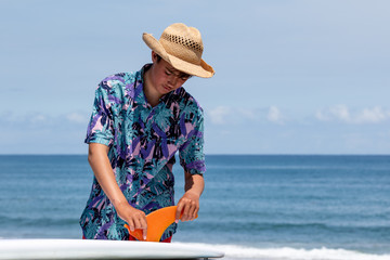 Putting a surfboard fin into a surfboard, the fin is orange in color and there is an Asian boy wearing a Hawaiian shirt and straw hat with red shorts. Chiba, Japan Surfing In Japan, Japan Surf