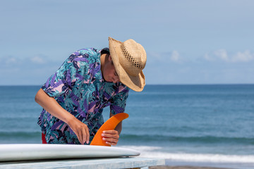 Fototapeta premium Putting a surfboard fin into a surfboard, the fin is orange in color and there is an Asian boy wearing a Hawaiian shirt and straw hat with red shorts. Chiba, Japan Surfing In Japan, Japan Surf