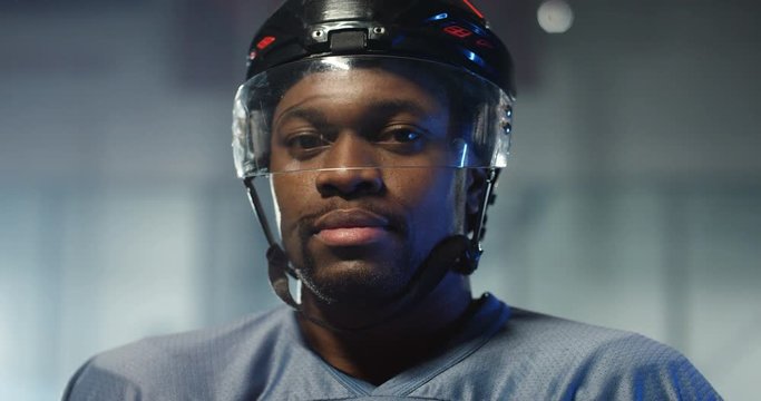Close up of happy handsome young African American male hockey player in helmet smiling cheerfully and breathing with steam on ice arena. Portrait of joyful sportsman.