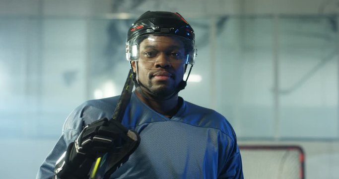 Portrait of happy handsome young African American male hockey player in helmet and with club smiling to camera on ice arena. Close up of cheerful attractive sportsman in casque with stick.