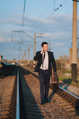 A cool and modern male businessman in a suit with a suitcase is walking along the paths from the train and talking on the phone. Business walk and serious conversation. Late for the train.