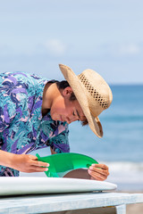 Fototapeta premium Surfer putting a green fin into a white surfboard on the beach with a blue ocean in the background he is Asian and wearing Hawaiian shirt and straw hat. He is also has a suntan