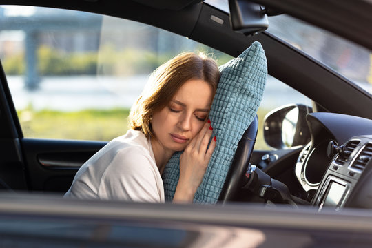 Tired young woman driver asleep on pillow on steering wheel, resting after long hours driving a car. Fatigue. Sleep deprivation. 