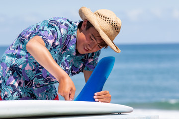 Fototapeta premium Japan Surf a boy putting a bluefin into a surfboard on the beach in Chiba close to the Olympic Surfing venue. He is wearing a Hawaiian shirt and straw hat & has a suntan. he is Japanese & has a smile.