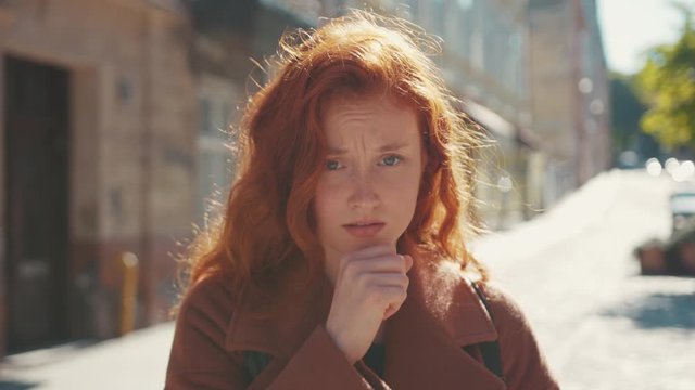 Poor unhealthy ginger woman with red curly hair standing on street sneezing allergy coughing suffering flu symptoms. Coronavirus. Virus infections. Pandemic in european city.