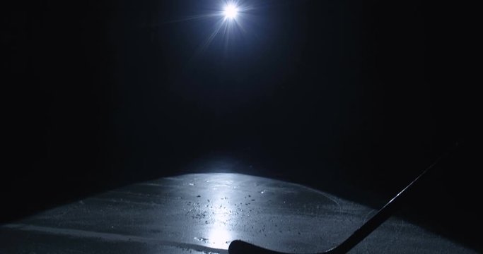 Close up of club hitting puck on icy surface in darkness. Hockey stick hits disc on ice arena in light of single spotlight lamp. Hockey match concept.