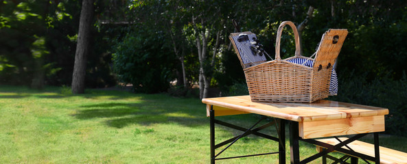 Open picnic basket on wooden table in green park, space for text. Banner design
