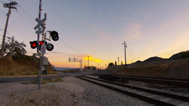 A railway crossing gate's lights flash as it opens after a train has passed and allows cars to go through at sunset.
