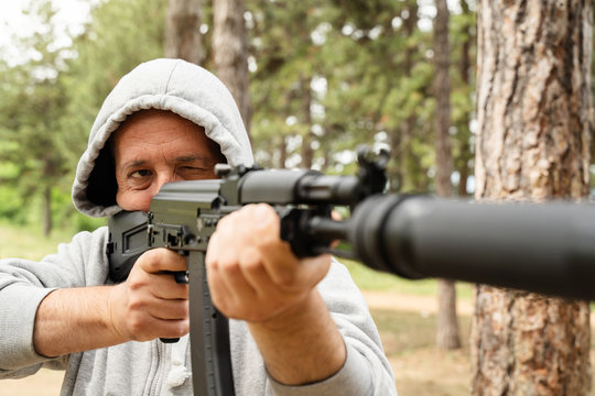 Front view of caucasian man wearing hood holding rifle semi automatic gun with silencer in the woods aiming ready to shoot or kill terrorist assassin murderer aim shooting in nature sunny day