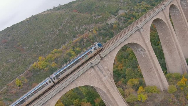 Aerial view closely following and diving around an SNCF passenger train crossing an old viaduct in a natural valley of France.