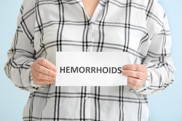 Elderly woman holding paper with text HEMORRHOIDS on color background