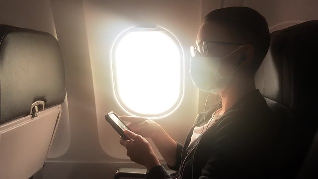 Young androgynous woman wearing disposable face mask plays on her phone and then sips from an iced coffee on a plane during the covid-19 pandemic