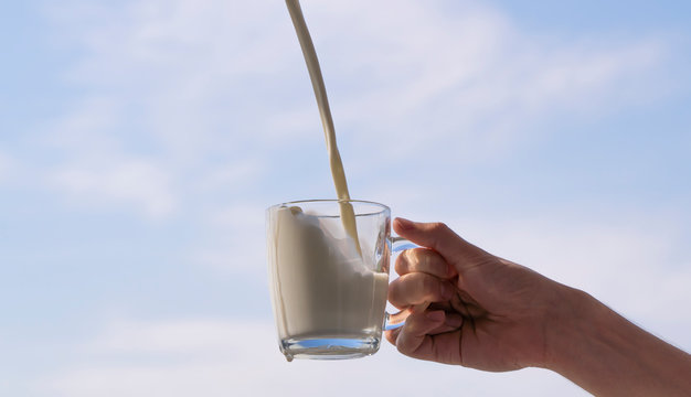 milk pours into the cup. hand holding a glass glass with dairy product against a clear sky