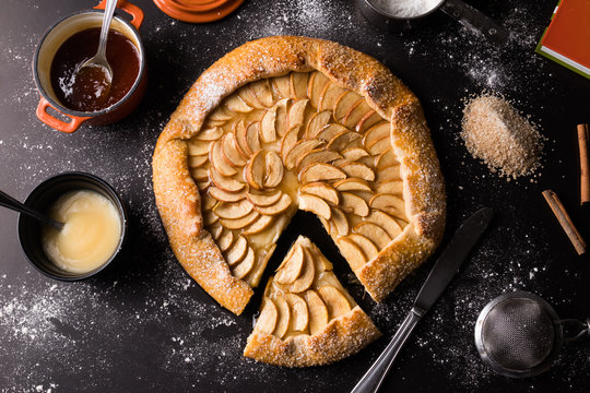 Pizza doce de creme com ma&ccedil;a e cobertura de caramelo sobre a mesa de cor preta com acess&oacute;rios - Borda a&ccedil;ucarada, ca&ccedil;arola laranja com caramelo