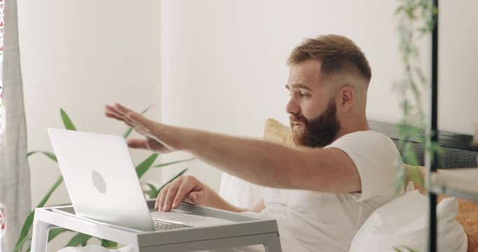 Side view of adult guy closing his laptop computer, gesturing and looking to window. Bearded man in 30s looking irritated and drinking juice while sitting on bed. Concept of bad news.
