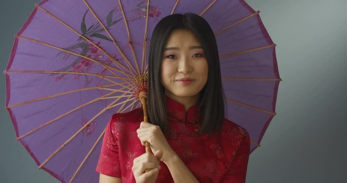 Portrait shot of Asian young beutiful woman in red cheongsam and with pink umbrella turning face to camera and smiling happily. Pretty girl in geisha style covered with parasol.