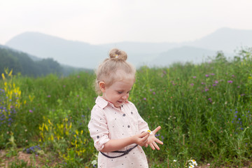 a blonde girl 2 years old in a dress stands in the middle of a flower meadow in the mountains and laughs