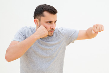 Serious young man standing in fighting pose with tightening fists and punching. Concentrated Caucasian guy training boxing and holding fist near face. Sport and fitness during quarantine concept