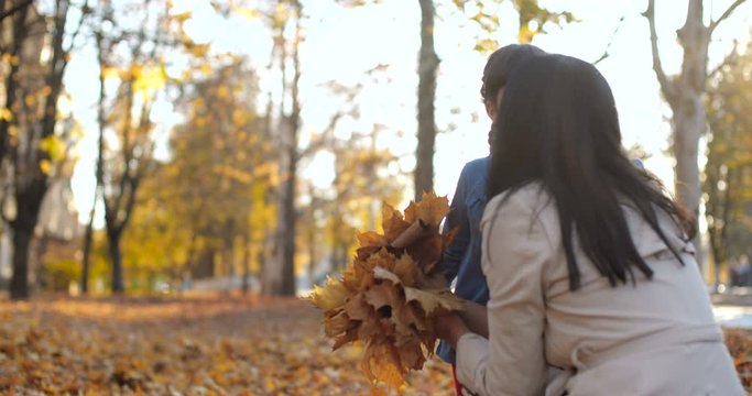 Caucasian brunette mother in white trench and her two daughters sit in city park. Girls collect fallen leaves and give bouquets to mom. Carpet of leaves. Positive emotions 50 fps slow motion