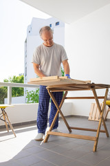 Mature man assembling wooden table on home balcony, doing DIY work. Vertical full length shot. Home interior improvement concept