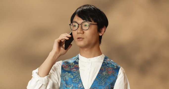 Young Asian handsome man in glasses and traditional clothes talking on mobile phone. Chinese good looking guy speaking on cellphone. Telephone call and conversation concept.