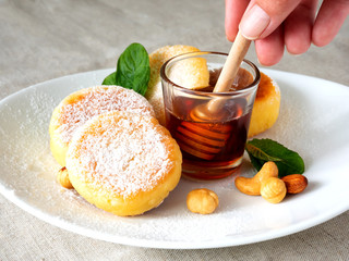Cheesecakes with fresh mint, nuts and honey. syrniki on a white plate. cottage cheese pancakes sprinkled with powdered sugar. Female hand holds a honey dipper