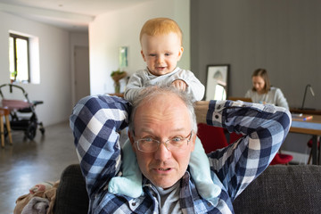 Cute baby sitting on grandfather shoulders and touching his hair. Senior man in glasses holding...