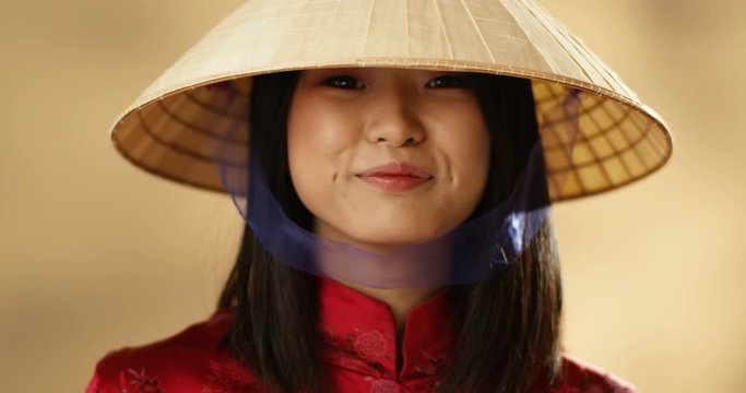 Close up of young Asian pretty happy woman in red clothes and conus hat rising face to camera and smiling cheerfully. Portrait of joyful beautiful girl in traditional female Chinese outfit with smile.