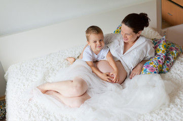 Obraz na płótnie Canvas Active pregnant woman sitting on bed and touching her belly at home, with her little son hugs belly. A pregnant smiling girl is resting with her family on a white bed. The last months of pregnancy.