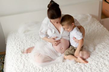 Active pregnant woman sitting on bed and touching her belly at home, with her little son hugs belly. A pregnant smiling girl is resting with her family on a white bed. The last months of pregnancy.