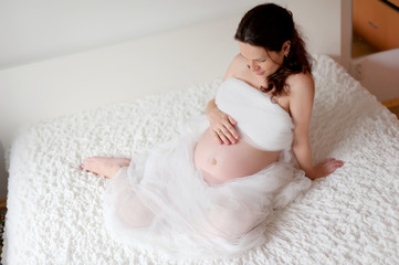 Active pregnant woman sitting on bed and touching her belly at home. A pregnant smiling girl is resting on a white bed. The last months of pregnancy.
