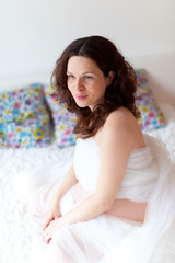 Obraz na płótnie Canvas Active pregnant woman sitting on bed and touching her belly at home. A pregnant smiling girl is resting on a white bed. The last months of pregnancy.