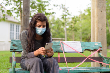 Young Indian woman with mask using phone while sitting with distance on park bench