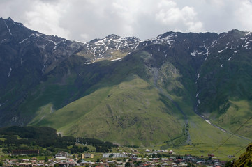 Fototapeta premium A huge mountain landscape towering above the gorge with a small town below. Snow-capped peaks of green hills covered with trees in the shade of clouds